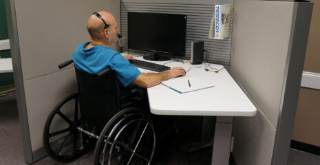 Man in a wheelchair sat at an office desk at a computer with a headset on.